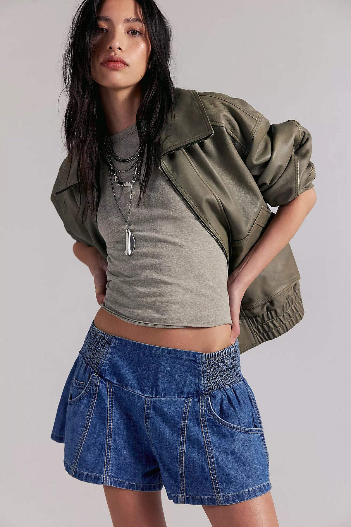 Woman wearing a green jacket, gray crop top, and blue shorts on a plain background
