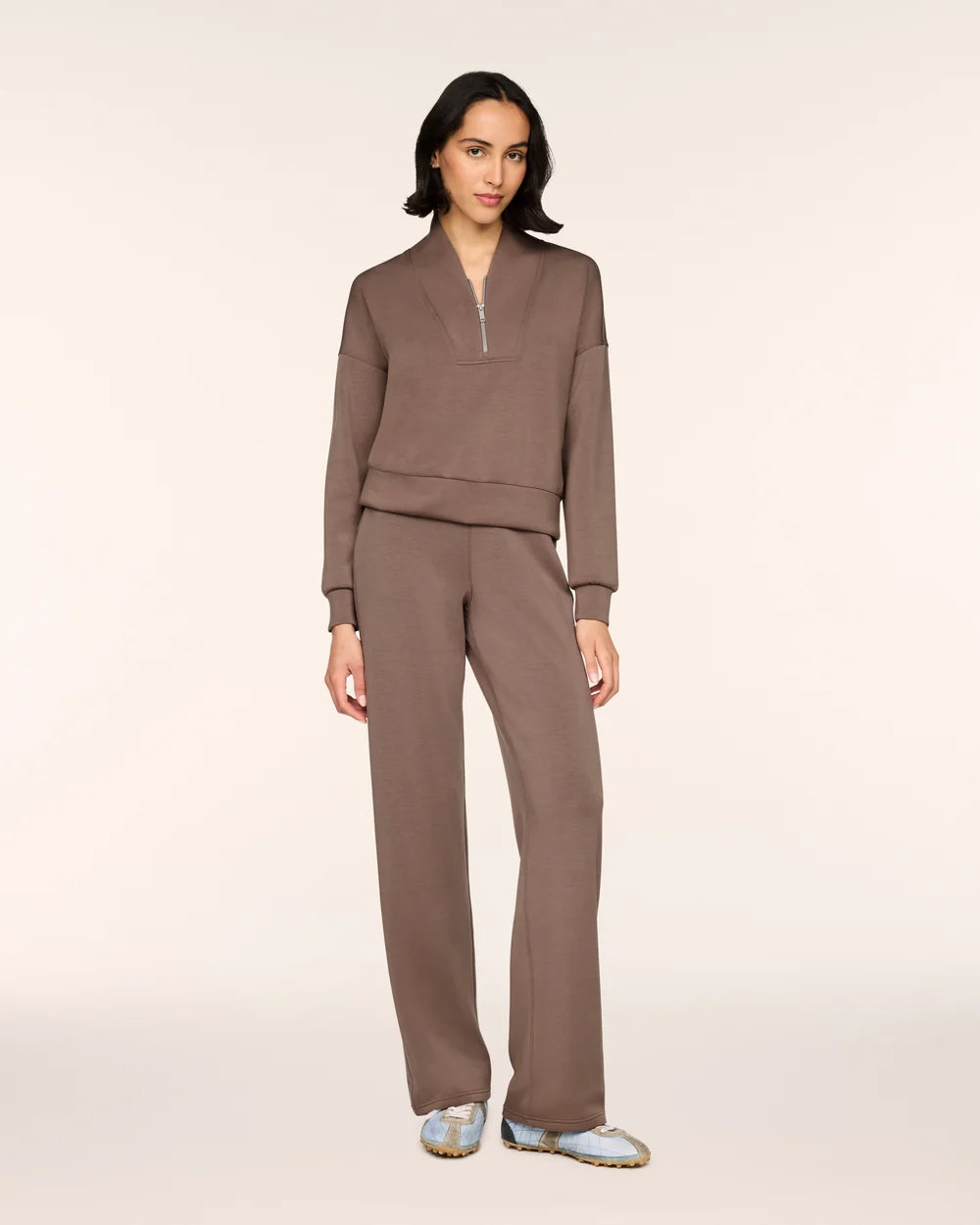A model stands against a plain light beige studio background wearing a matching taupe-brown lounge set consisting of a long-sleeve half-zip pullover and wide-leg pants, paired with light blue and white sneakers.