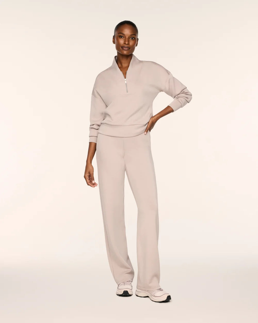 A model wearing a light beige two-piece tracksuit consisting of a long-sleeve quarter-zip pullover and matching wide-leg sweatpants, paired with white sneakers, against a solid off-white studio background.