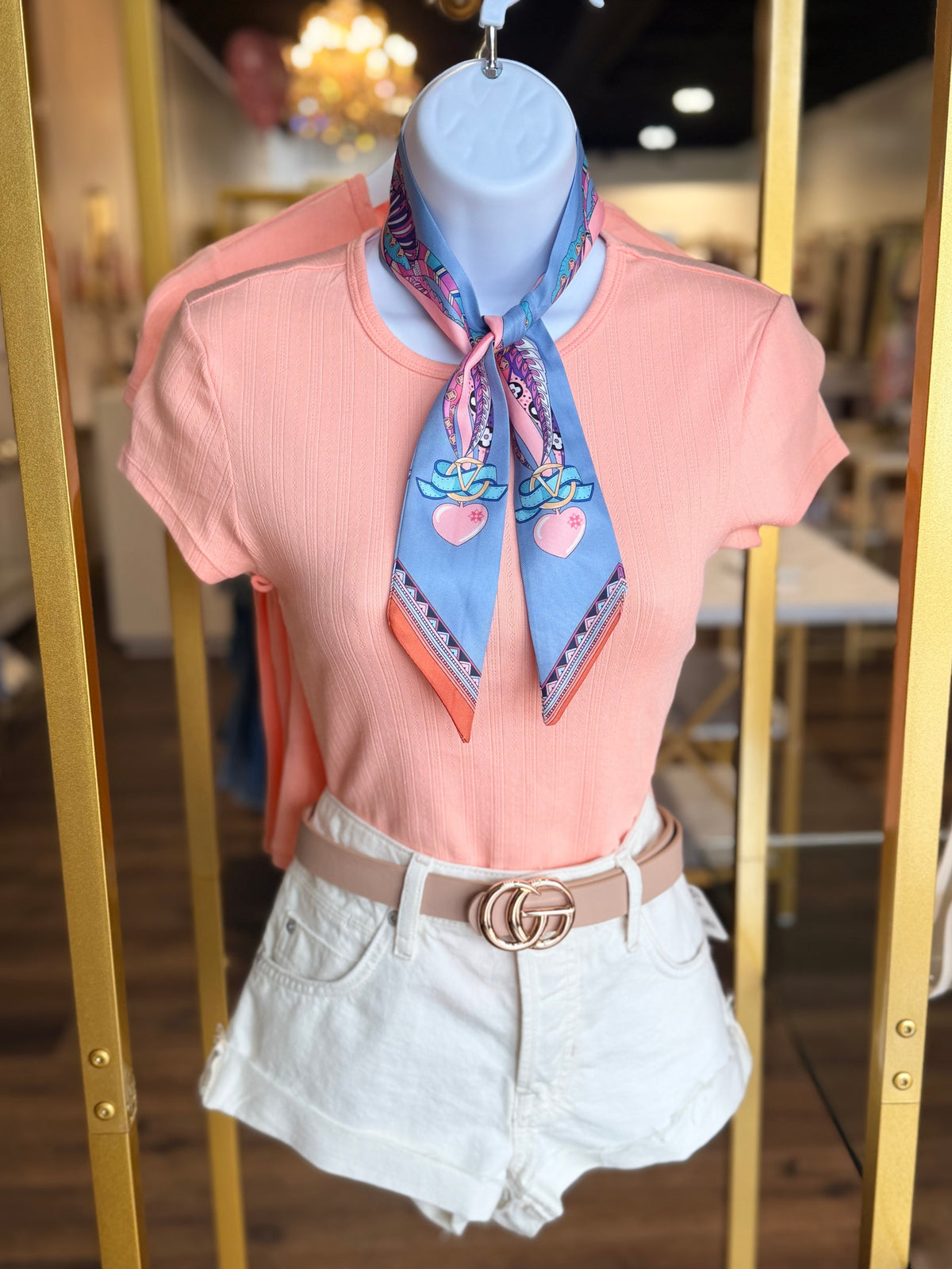 Mannequin wearing a pink top, white shorts, and a colorful scarf with a blurred indoor background.