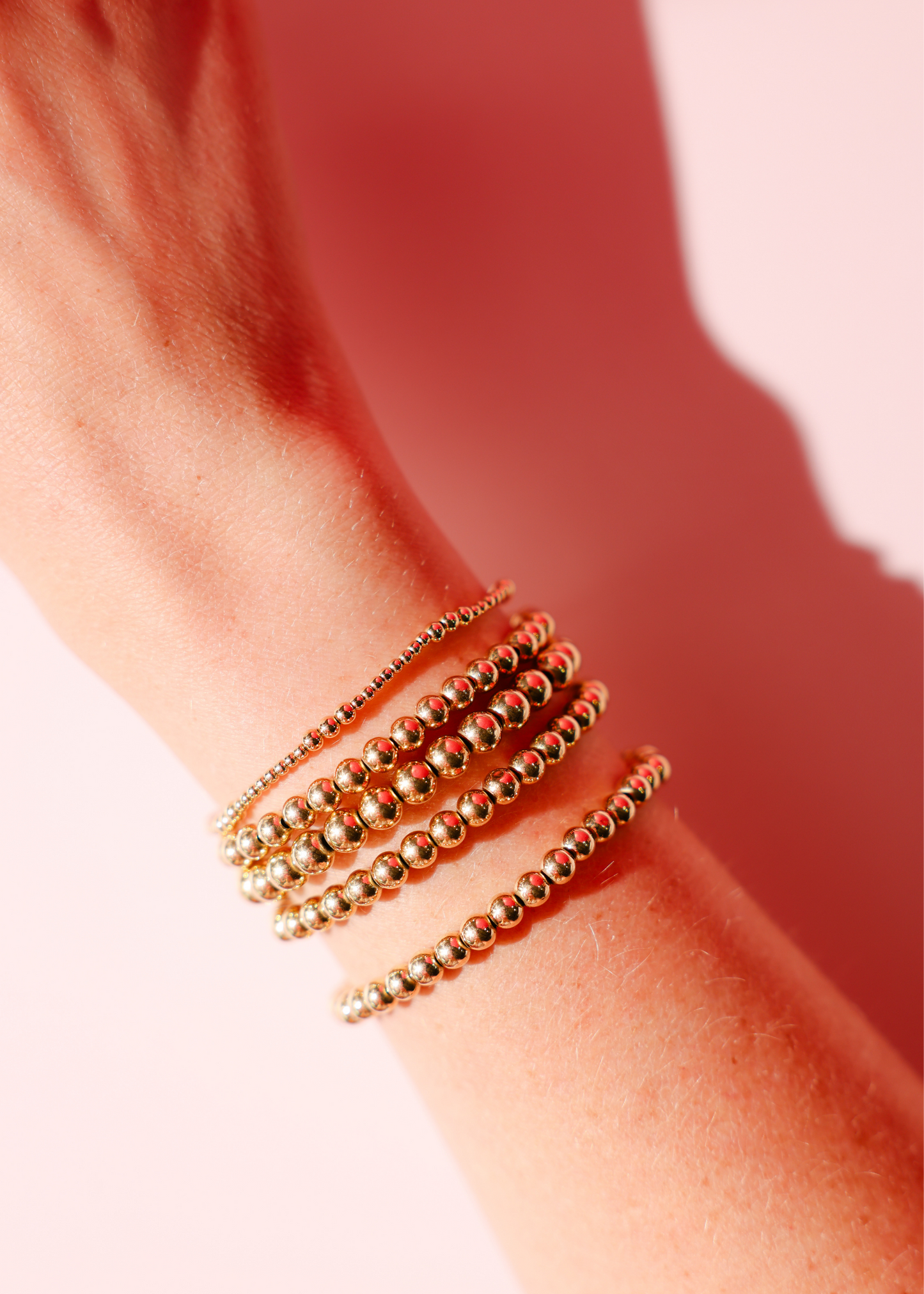 Close-up of a wrist wearing multiple gold beaded bracelets on a pink background