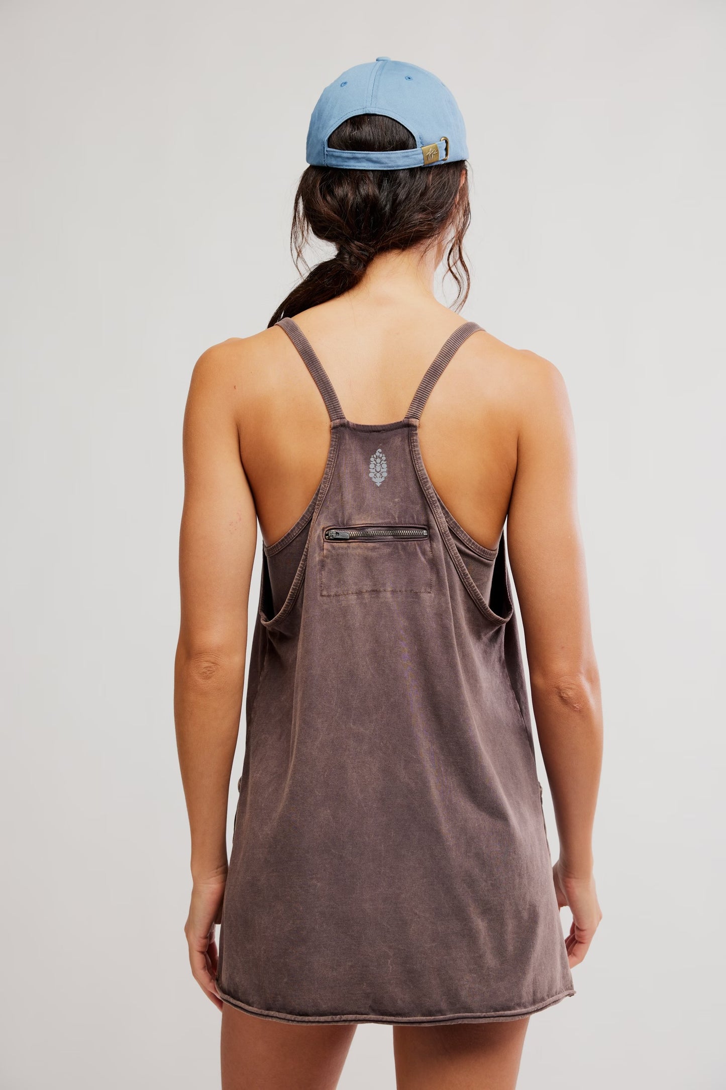 Person wearing a brown tank dress with a blue cap on a plain background