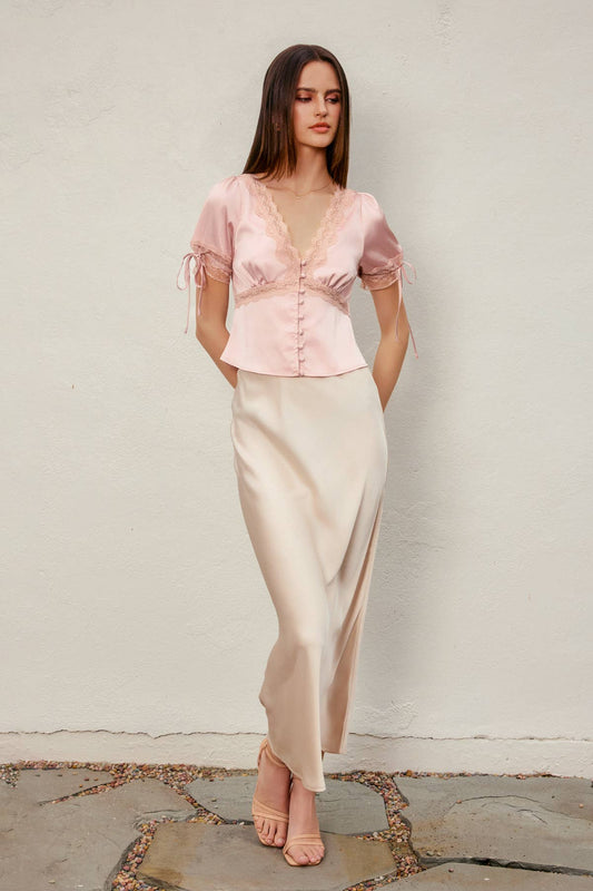 A woman modeling a light pink satin short-sleeve top with lace trim and a matching satin maxi skirt in a pale beige color, standing against a textured off-white wall.