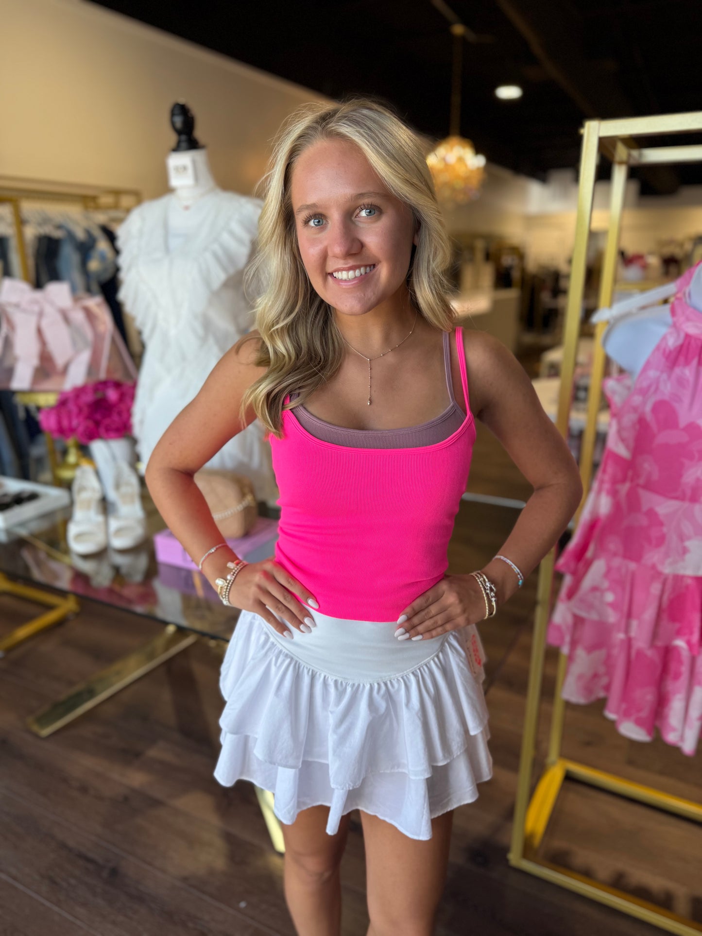 A smiling blonde woman stands in a boutique wearing a bright pink tank top and a white ruffled mini skirt. Clothing racks, mannequins, and store fixtures are visible in the background.