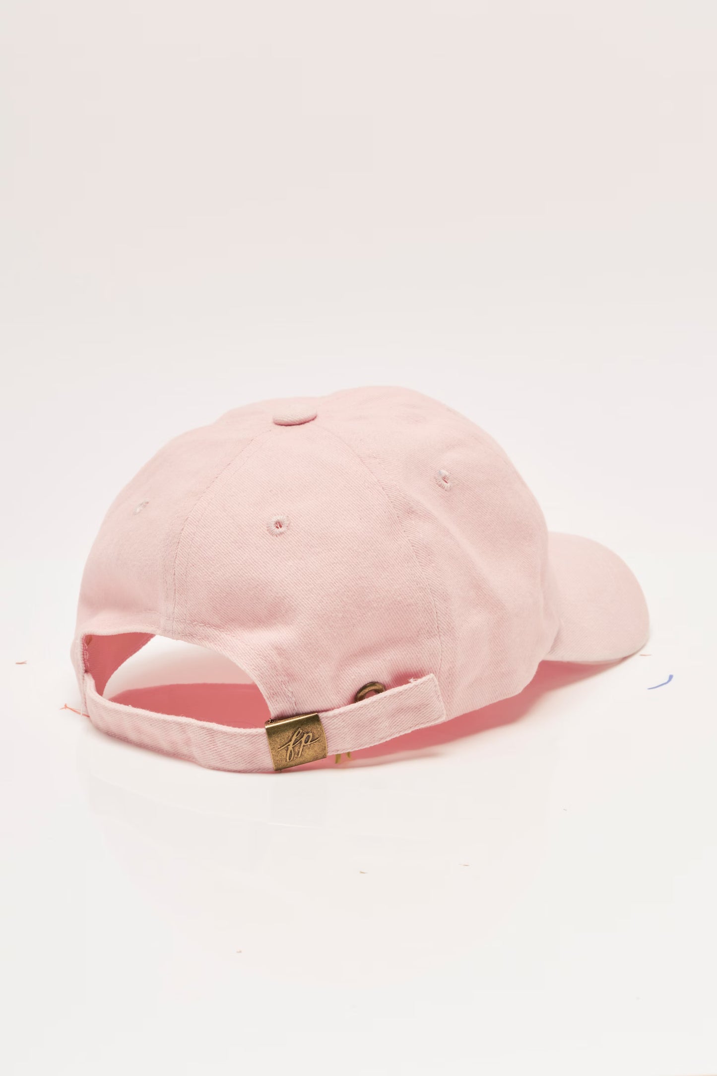 A light pink baseball cap shown from the back, featuring a matching pink adjustable strap with a small gold metal brand tag.