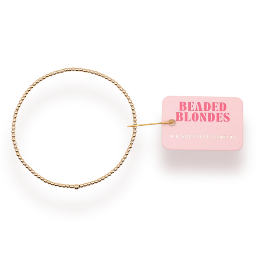 Beaded Blondes 2mm Gold Bracelet arranged in a circle beside a pink brand tag.