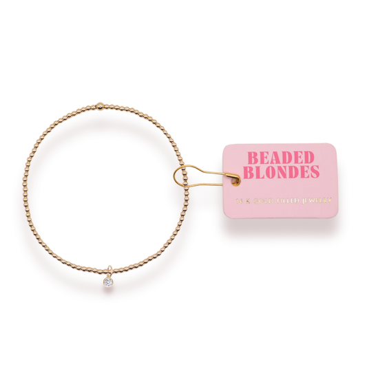 April Birthstone Charm Bracelet with a clear charm and gold beads beside a pink Beaded Blondes tag.