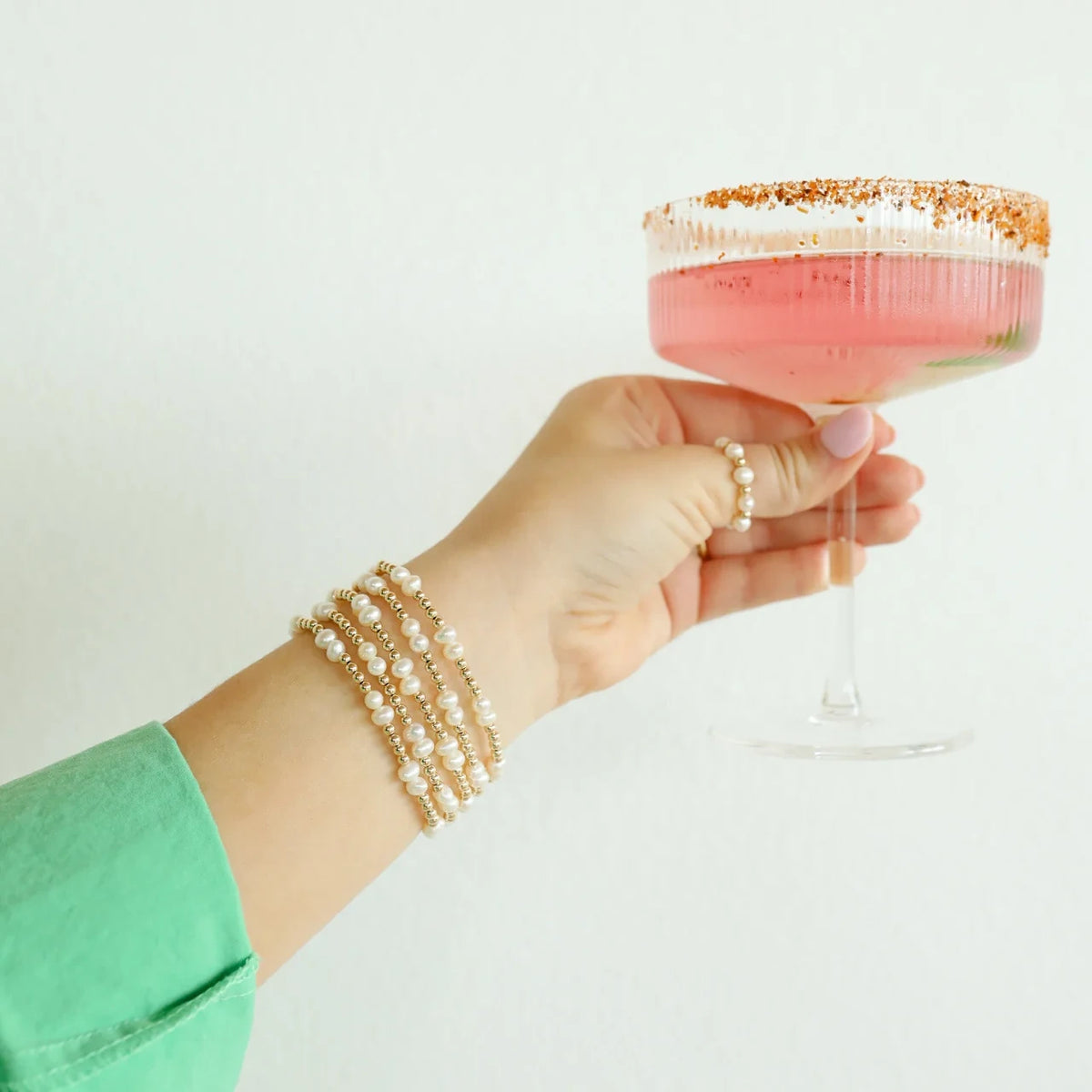 A hand wearing a pearl ring and stacked gold bracelets holds a pink cocktail glass.