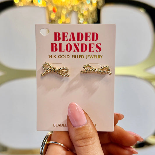 Gold beaded bow earrings on a Beaded Blondes card held in front of a boutique display.