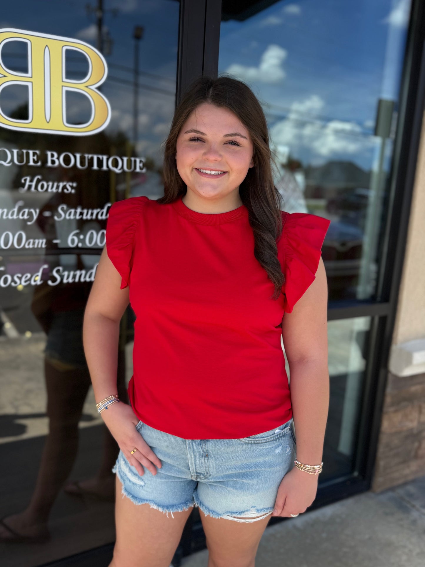 A smiling woman stands in front of a boutique glass door wearing a bright red ruffled-sleeve top and light blue denim shorts. The door has a gold and white 'BB Boutique Boutique' logo and business hours printed on it.
