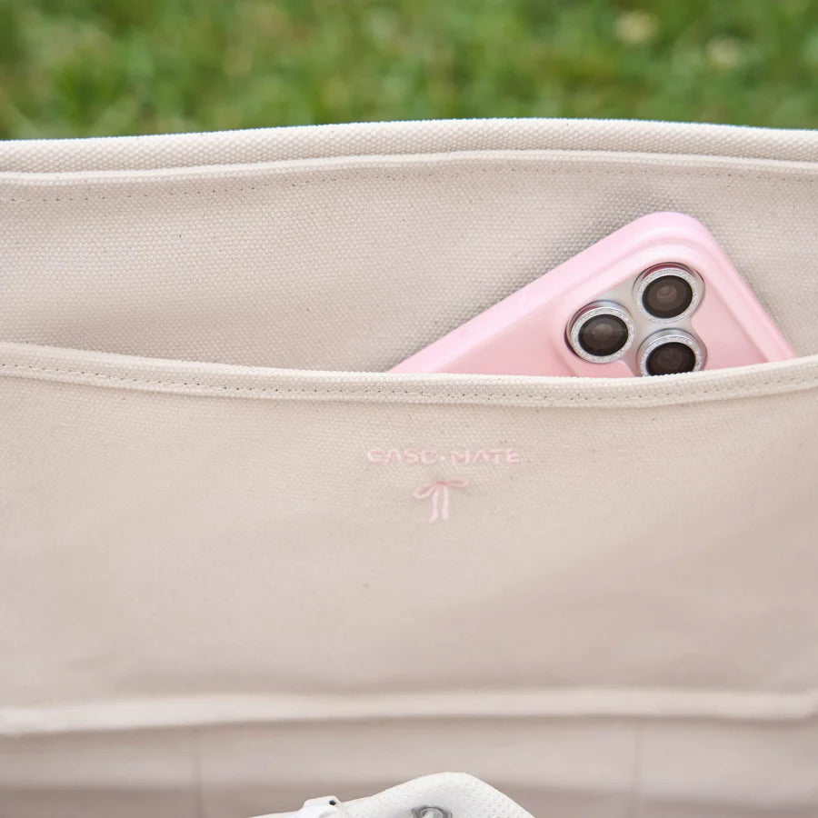 Close-up of a beige canvas shoulder bag with a pink smartphone tucked into the side pocket. The brand name CASLANATE is subtly embroidered on the front.