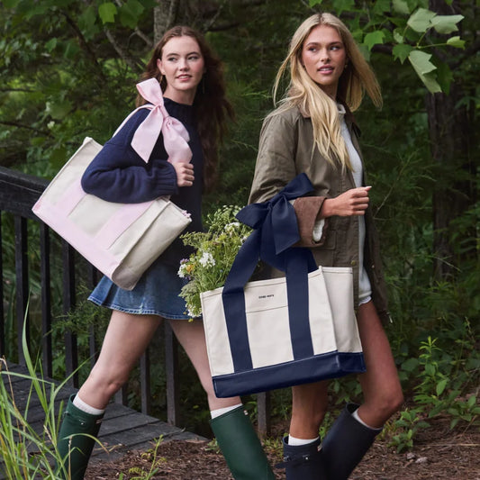 Two women walking outdoors in a wooded setting, each carrying a large canvas tote bag with a wide ribbon bow. The tote bags are cream with navy blue panels and handles.