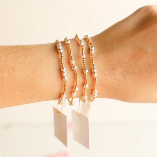 Three delicate gold beaded bracelets with small white pearl-like beads, worn stacked on a person's wrist against a soft off-white background.