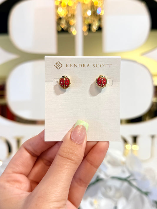 Red ladybug earrings on a Kendra Scott card held by a hand with a blurred festive background.