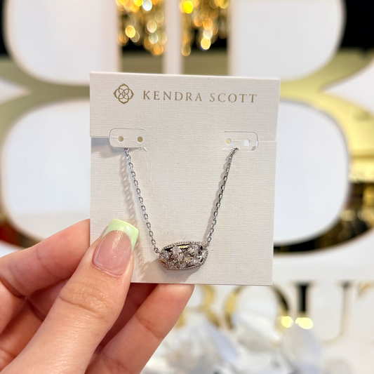 Hand holding a Kendra Scott necklace card with a blurred decorative background