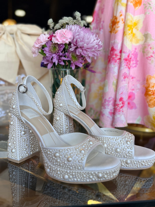 White pearl embellished high-heeled shoes on a reflective surface with flowers and a floral dress in the background.