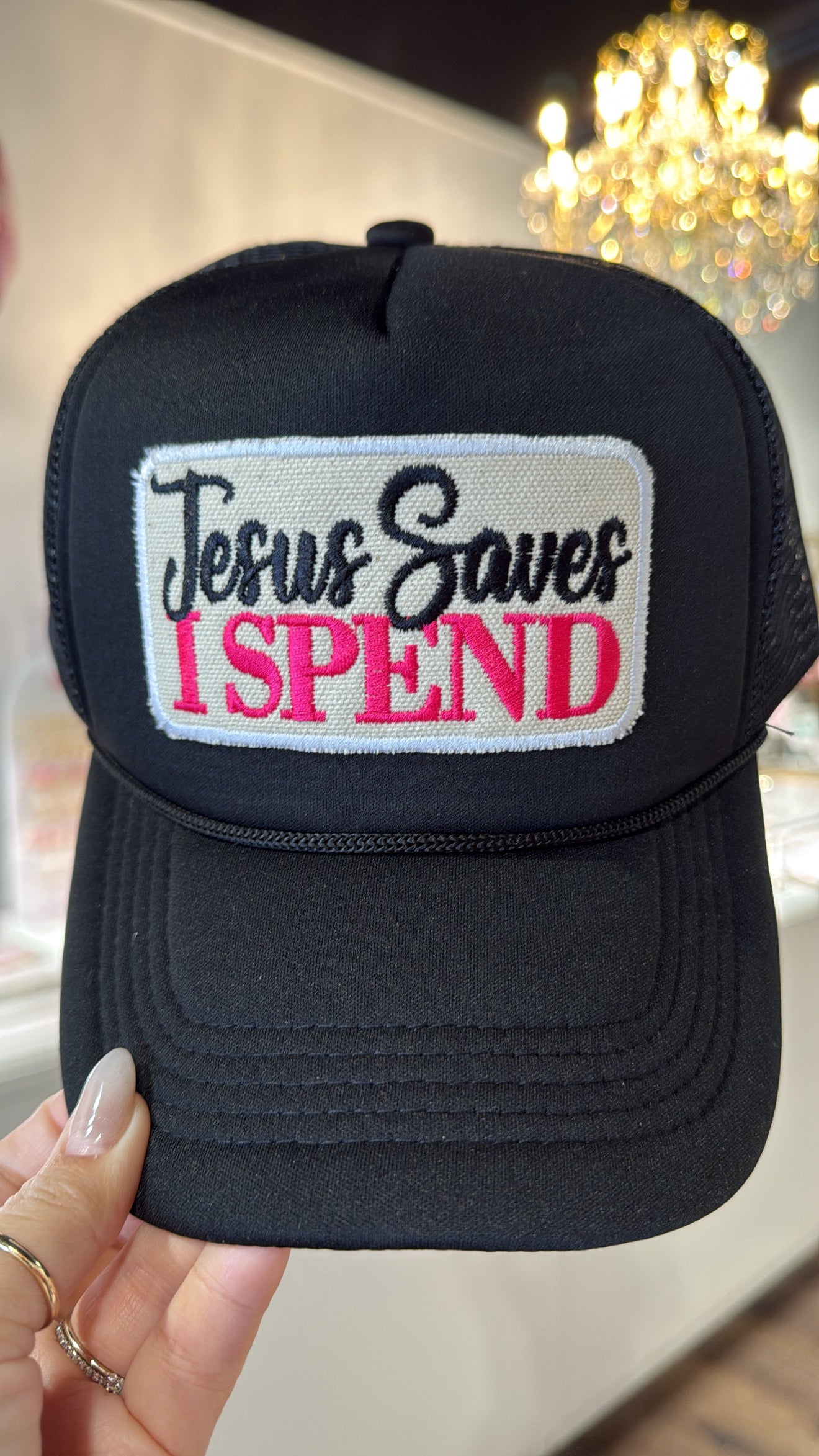 Black cap with 'Jesus Saves I Spend' text held by a hand.
