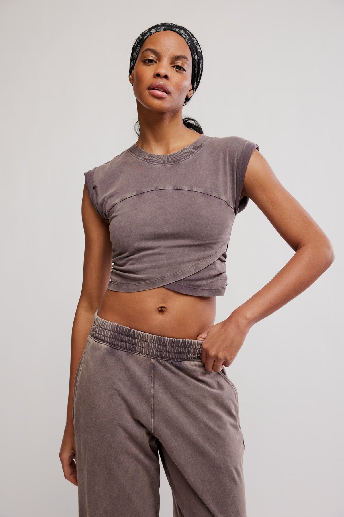 A woman modeling a cropped, sleeveless athletic top and matching jogger pants in a washed taupe-gray color, set against a plain light gray studio background.
