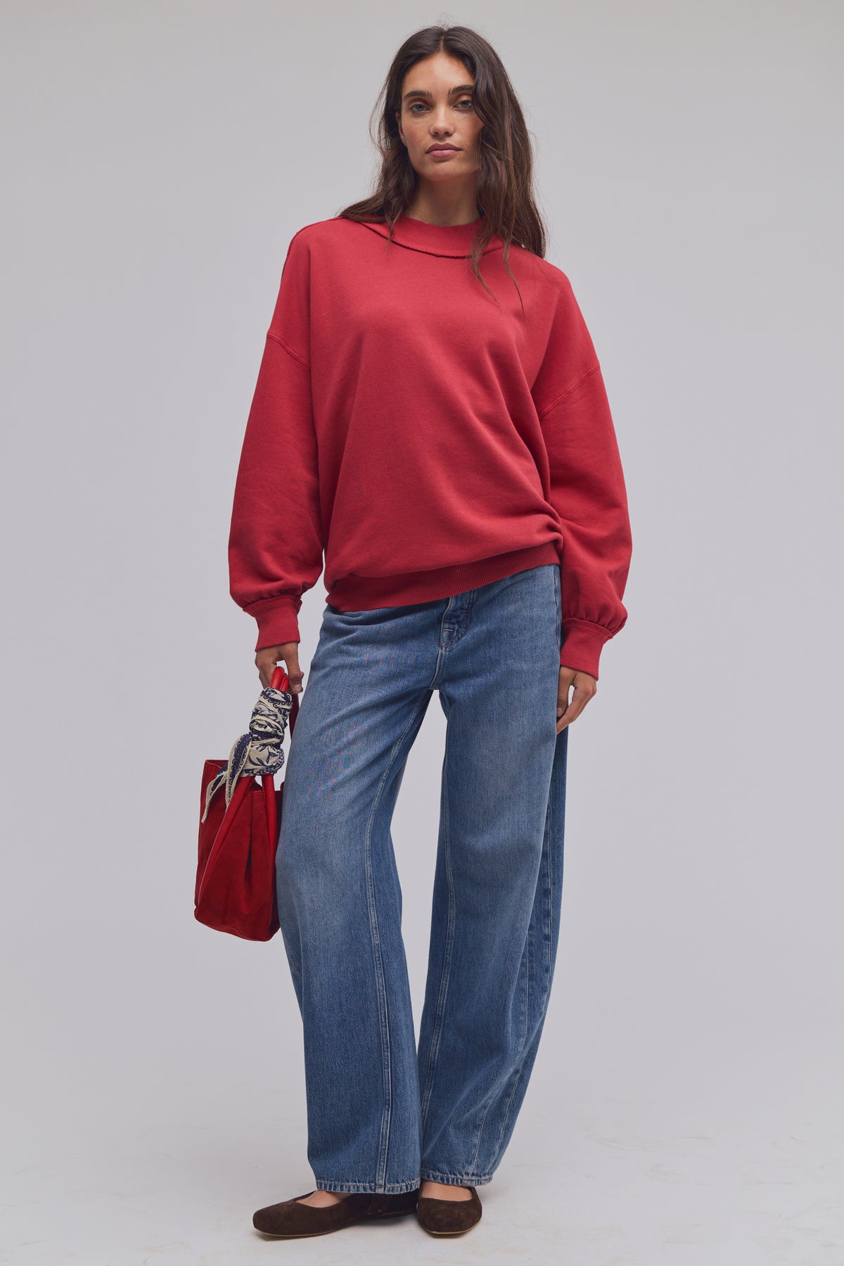 Person wearing a red sweater and blue jeans holding a red bag on a plain background