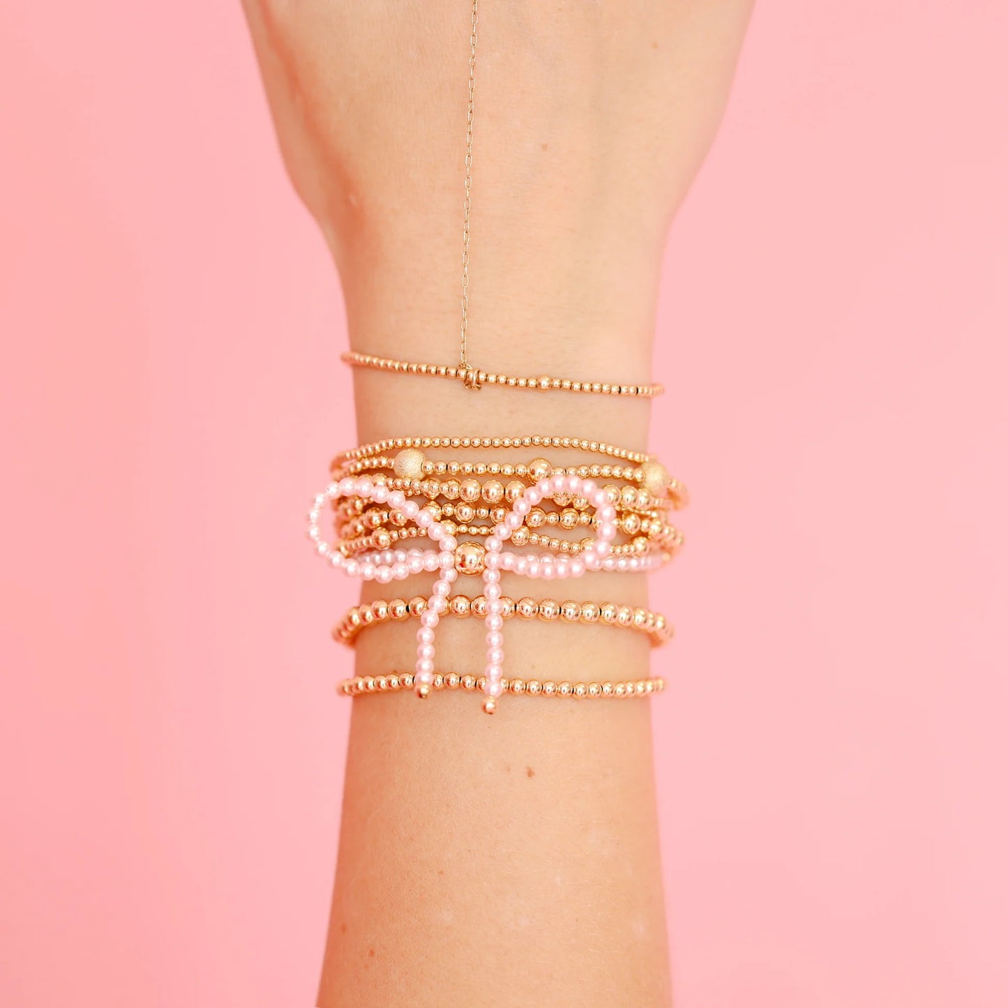 A stack of delicate beaded bracelets on a person's wrist, featuring gold-toned bead strands, white pearl-like beads, and a bow-shaped charm, set against a solid pink background.