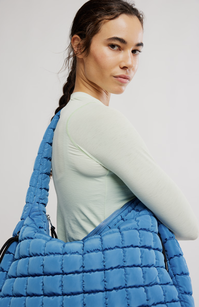 A woman wearing a pale mint green long-sleeve top carries a large quilted blue puffer shoulder bag against a light gray studio background.
