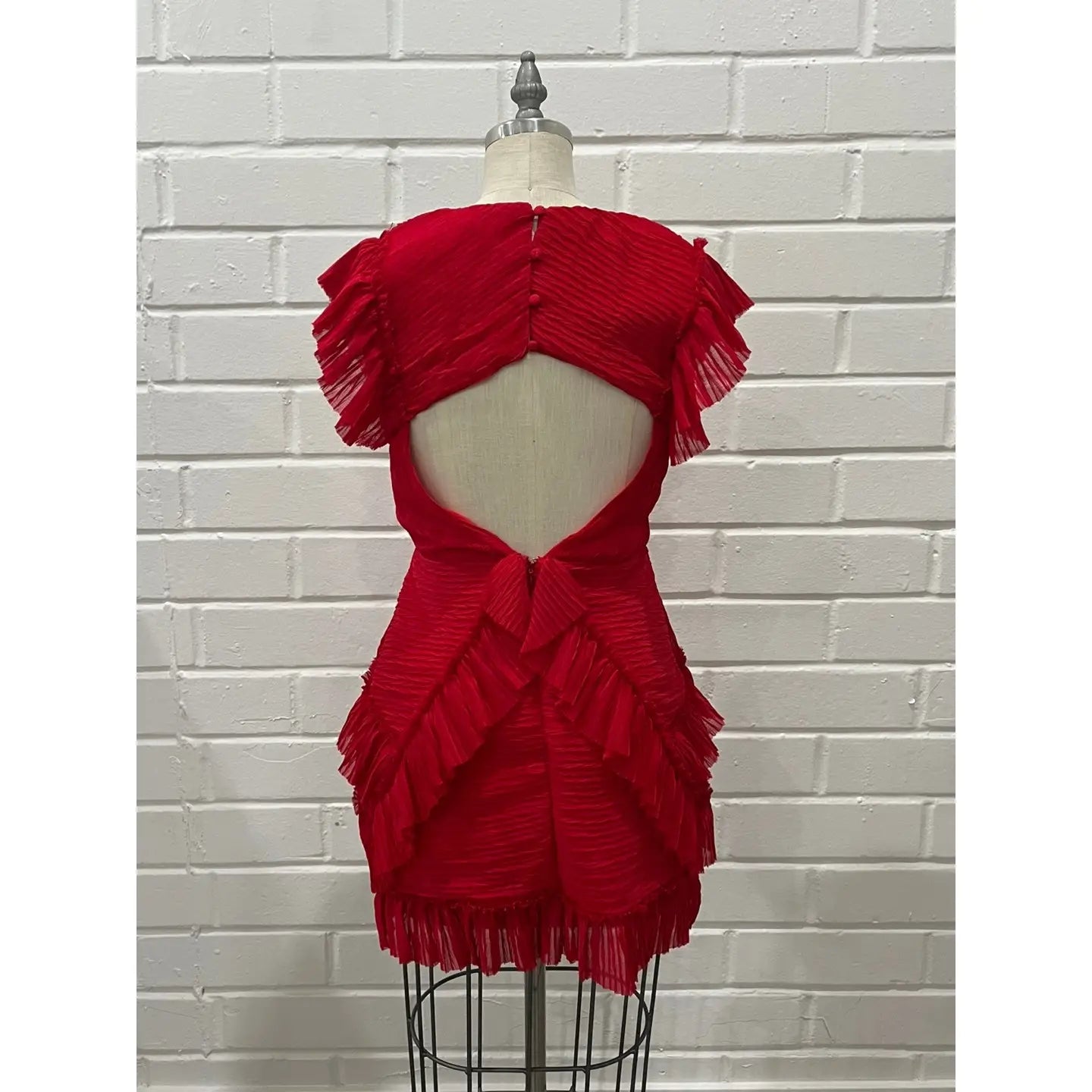 A bright red pleated mini dress with a dramatic cutout back and layered ruffle details, displayed on a dress form mannequin against a white brick wall background.