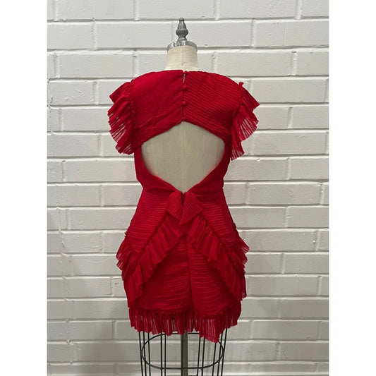 A bright red pleated mini dress with a dramatic cutout back and layered ruffle details, displayed on a dress form mannequin against a white brick wall background.