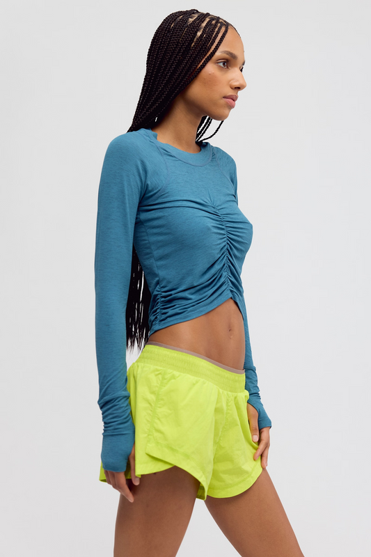 A woman modeling a fitted long-sleeve athletic crop top in teal blue with ruching and a side cutout, paired with neon yellow athletic shorts, against a light gray studio background.