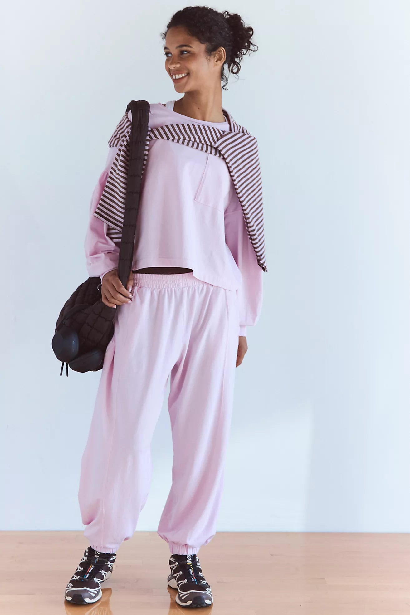 A woman modeling a light pink lounge set consisting of a cropped long-sleeve top with a black-and-white striped draped overlay and matching loose jogger pants, carrying a black shoulder bag.