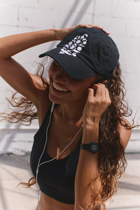 Woman wearing a black cap with a white logo, black top, and earphones, standing against a light-colored wall.