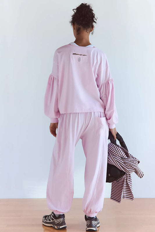 A model shown from behind wearing a light pink two-piece lounge set consisting of a long-sleeve sweatshirt with balloon sleeves and matching jogger pants. The model is holding a striped shirt and a black bag against a clean white wall and light wood floor.