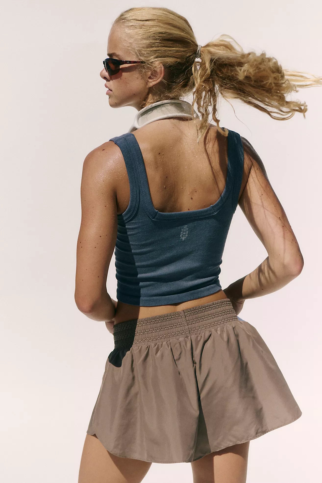 A woman shown from the back wearing a dark blue ribbed scoop-neck athletic tank top and a taupe-brown pleated athletic skirt, set against a plain light background.