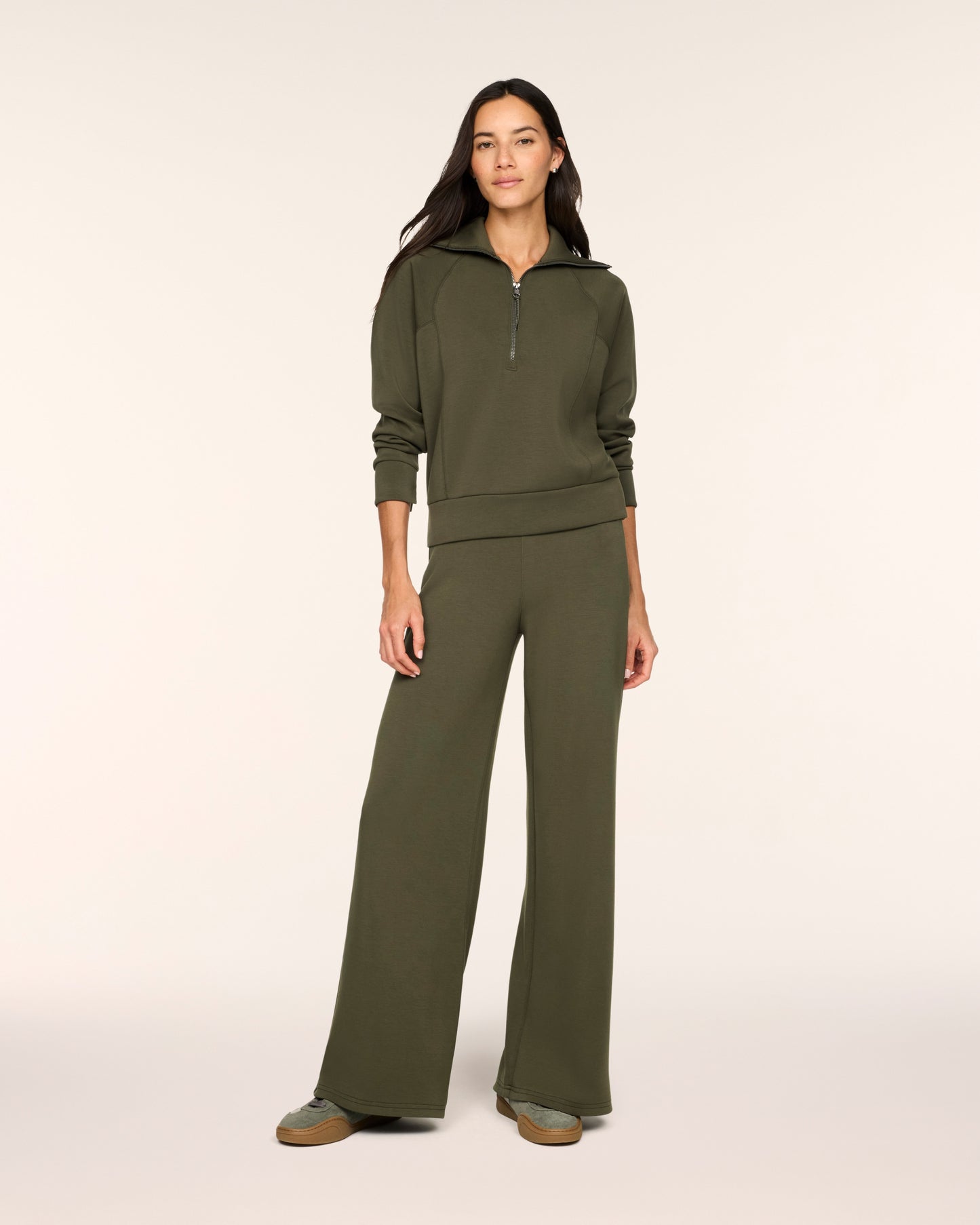 A woman modeling a matching olive green lounge set consisting of a half-zip long-sleeve top and wide-leg pants, photographed against a solid off-white studio background.