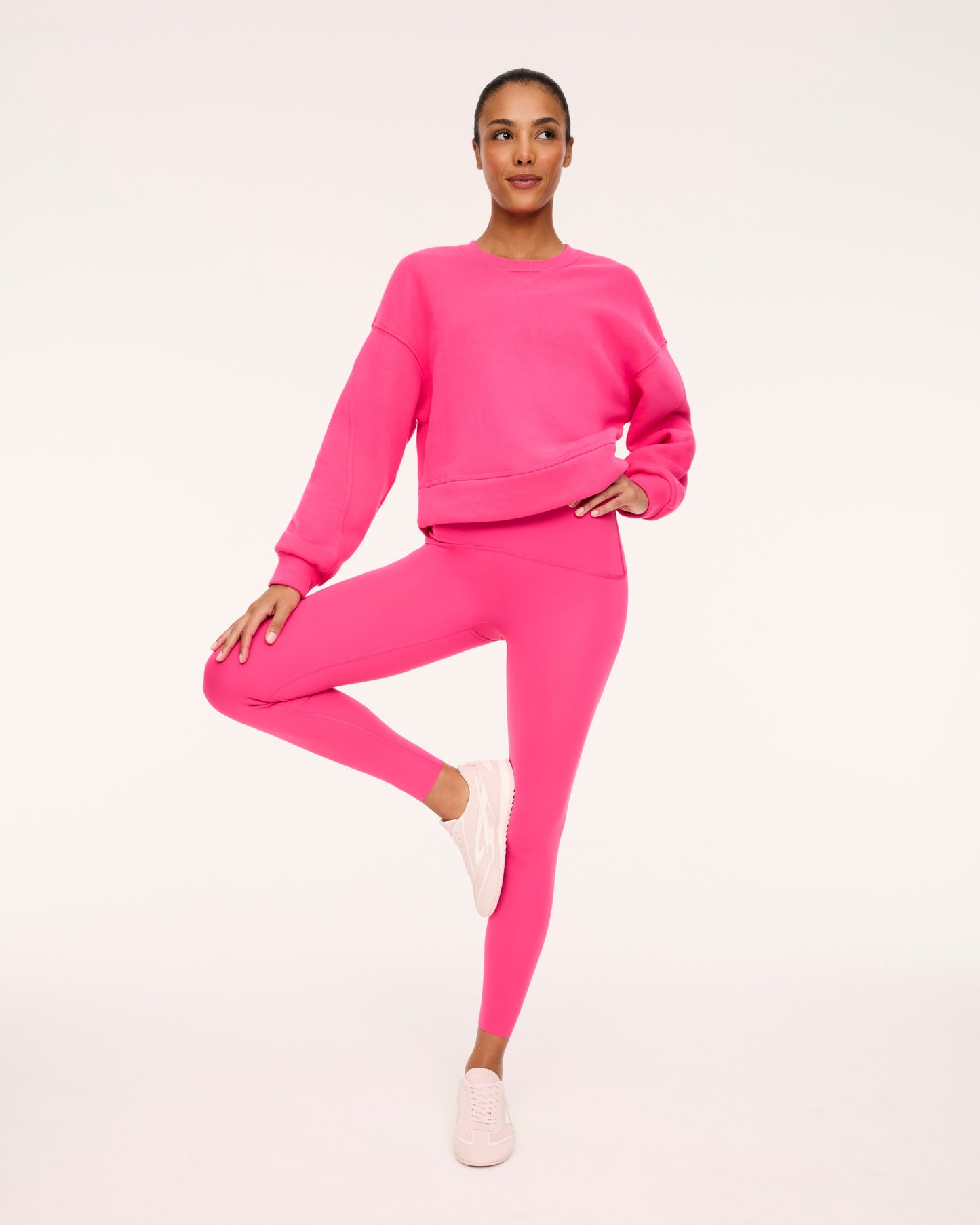 A model posing in a matching hot pink activewear set consisting of a cropped long-sleeve sweatshirt and high-waisted leggings, paired with light pink sneakers, against a solid off-white studio background.