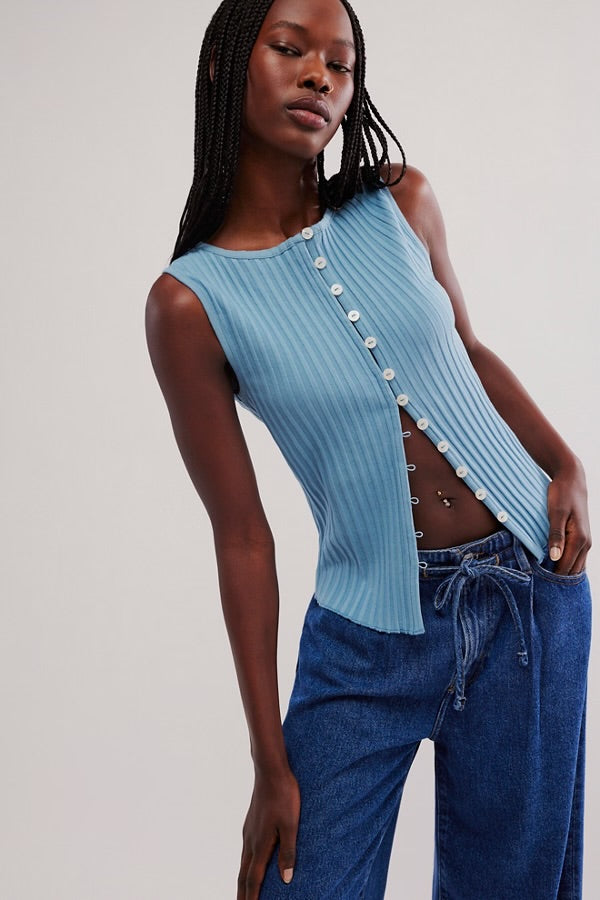 Woman wearing a light blue ribbed tank top with button details and blue jeans on a plain background