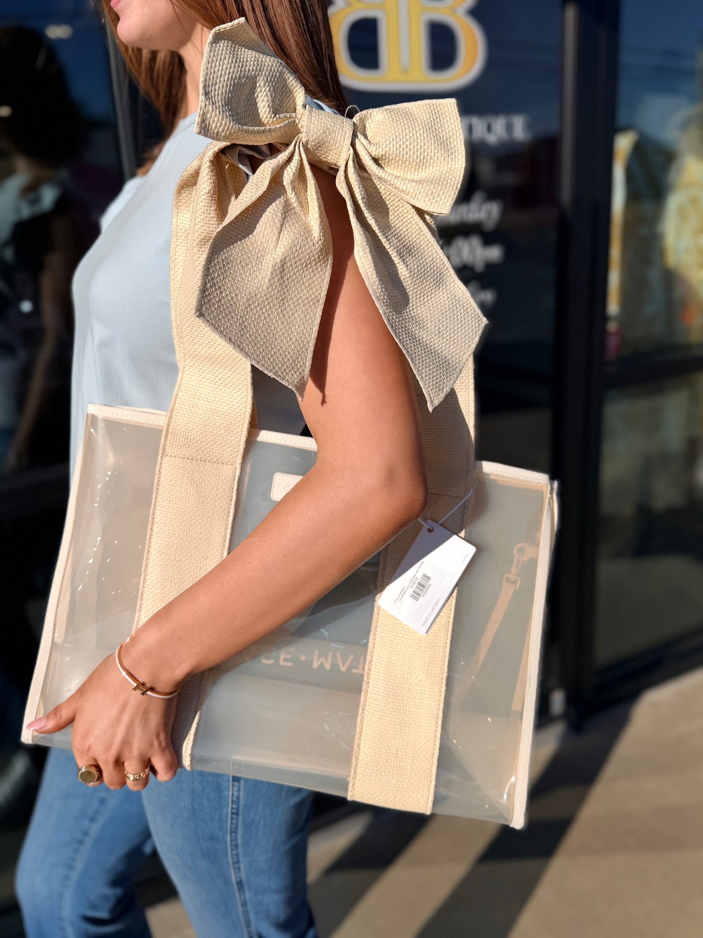 Person wearing a beige vest with a large bow, holding a transparent bag with a visible brand tag.