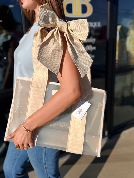 Person wearing a beige vest with a large bow, holding a transparent bag with a visible brand tag.