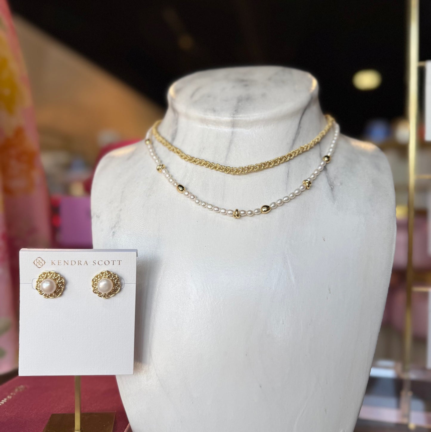 Three Kendra Scott jewelry pieces in front of store. Two necklaces on a bust and a pair of gold pearl earrings 