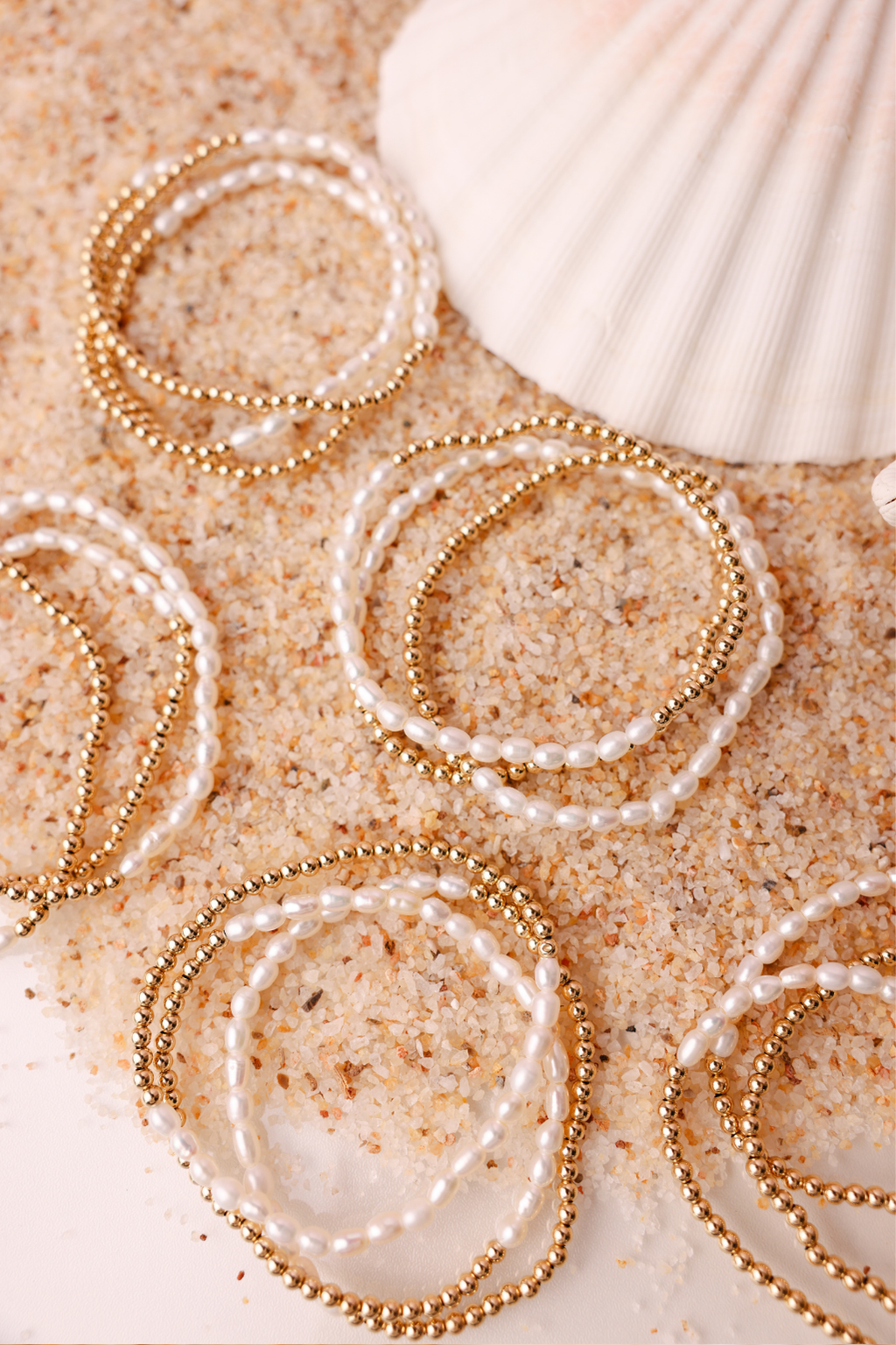 Gold and pearl bracelets on sand with a shell in the background