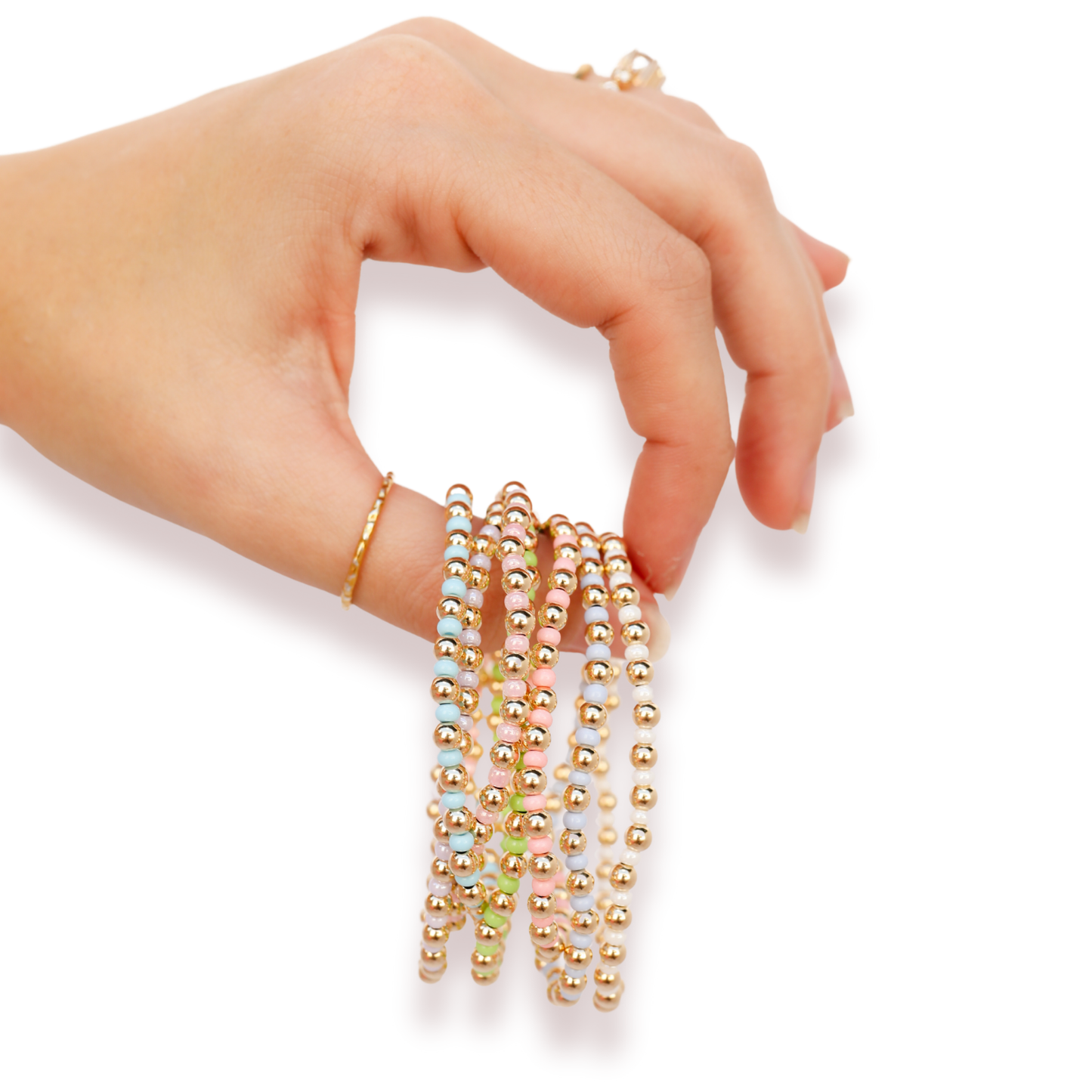 A hand wearing a delicate gold ring and holding a stack of multiple beaded strands in gold and iridescent pastel colors against a white background.