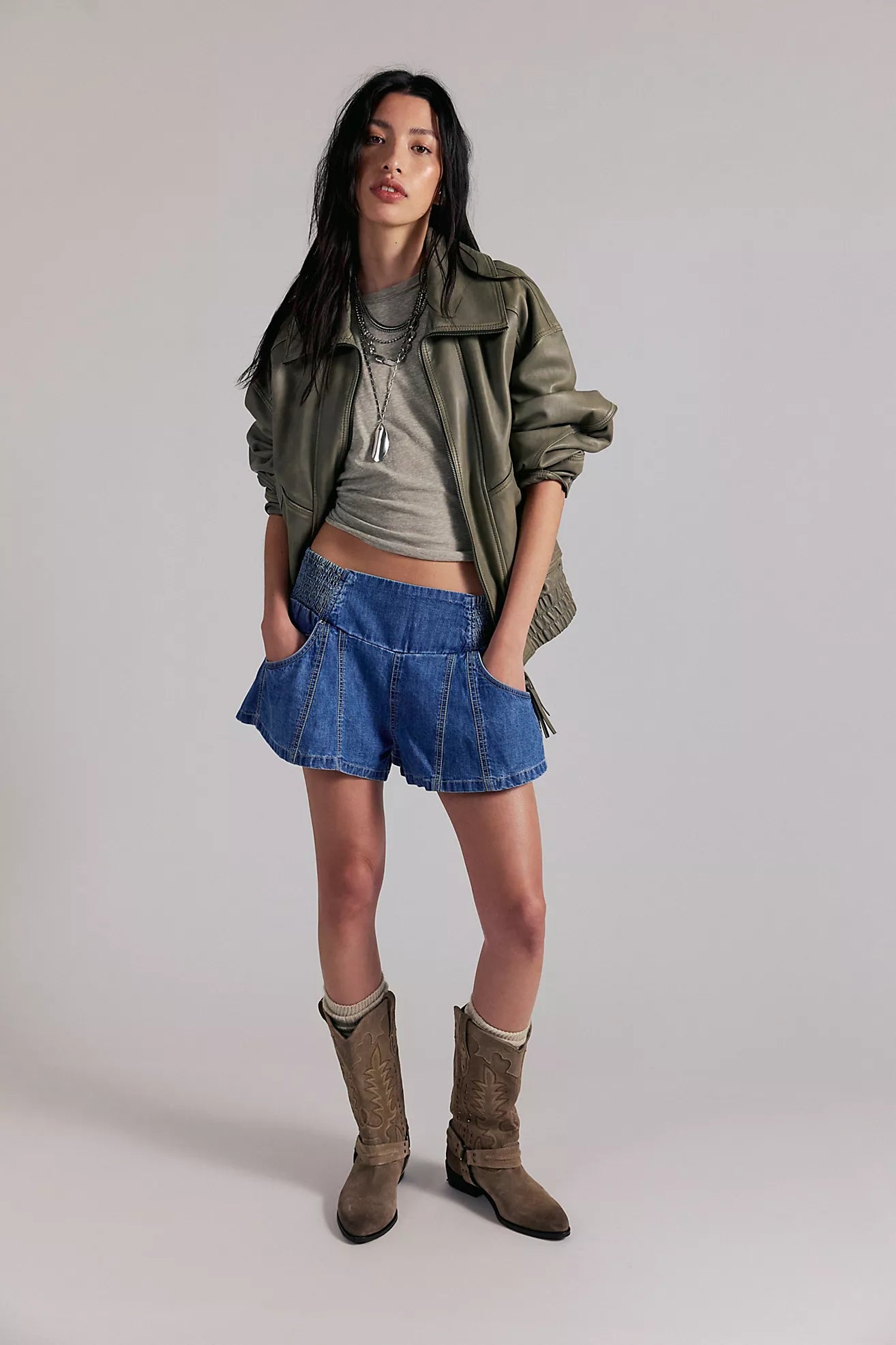 Woman wearing a green jacket, gray top, blue shorts, and brown boots on a plain background