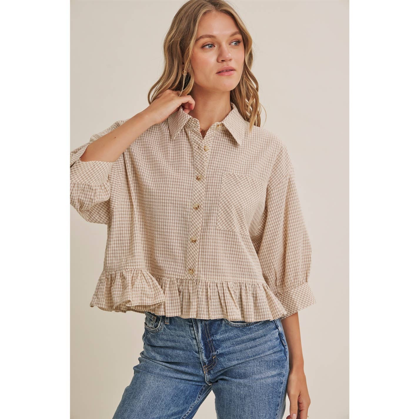 Woman wearing a checkered blouse with ruffled sleeves and blue jeans on a beige background