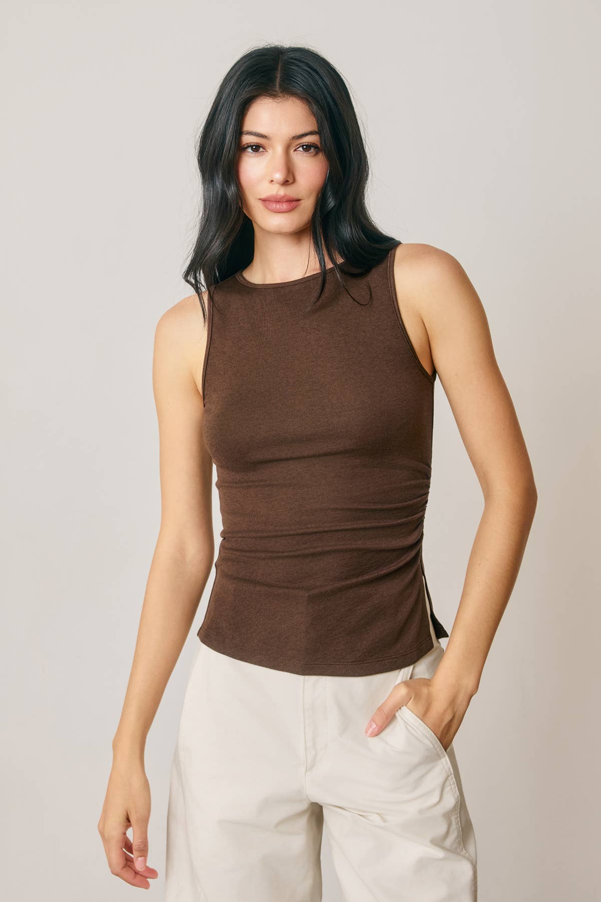 A woman modeling a sleeveless brown ribbed tank top with gathered ruching at the waist, paired with off-white pants, against a plain light gray studio background.