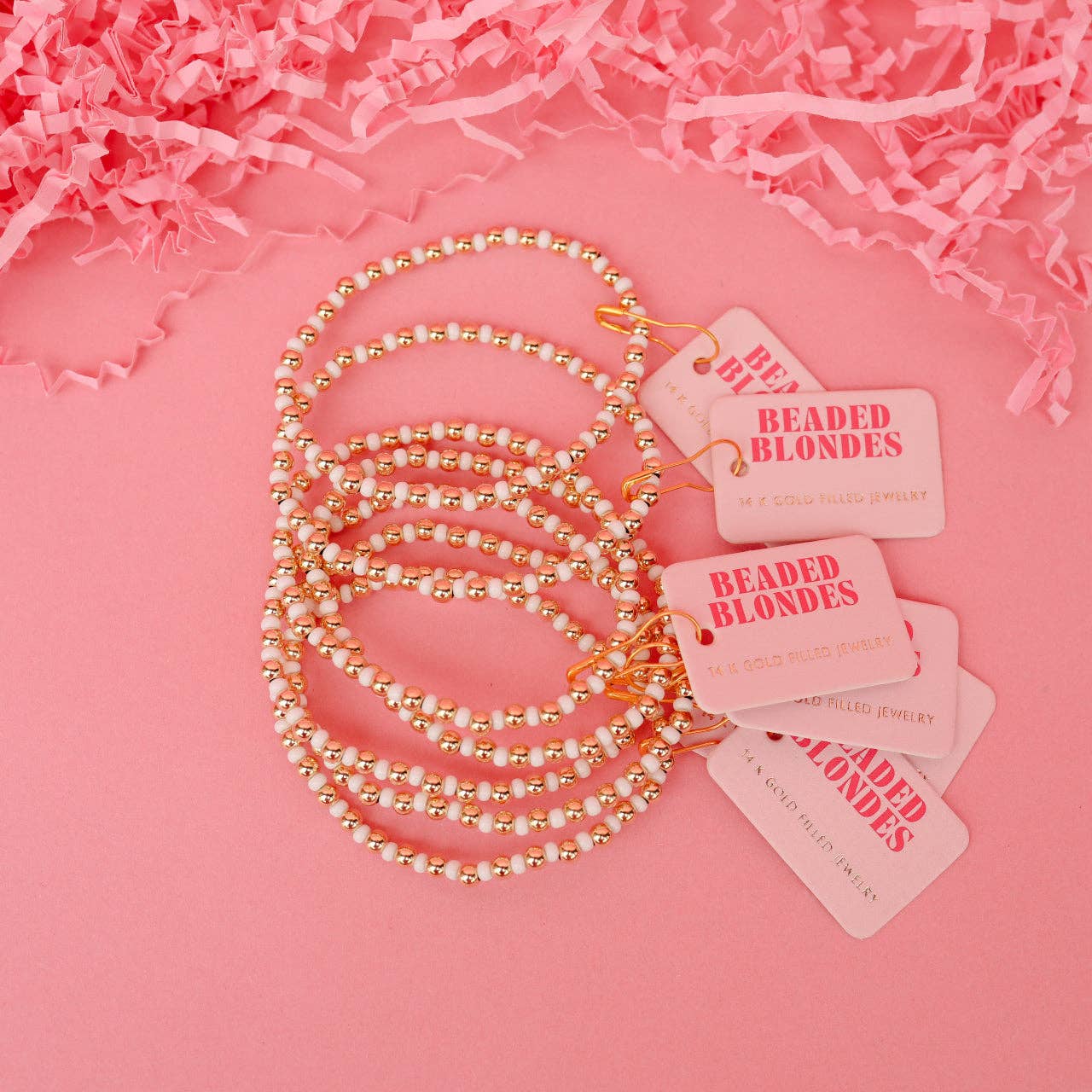 A set of five coiled beaded bracelets with gold-toned beads and white pearl-like beads, each attached to a pink tag reading 'BEADED BLONDE' on a pink background with shredded pink paper.