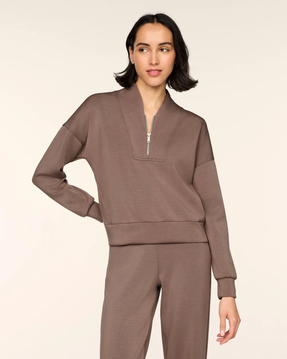 A woman modeling a matching taupe-brown lounge set consisting of a long-sleeve half-zip sweatshirt and coordinating sweatpants, shot against a solid light beige background.