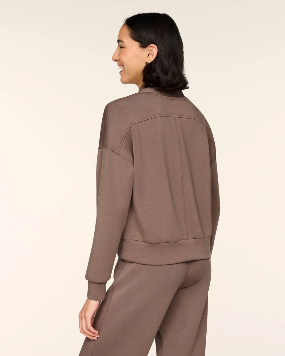 A woman shown from the back wearing a matching taupe-brown cropped sweatshirt and sweatpants set against a plain light beige studio background.