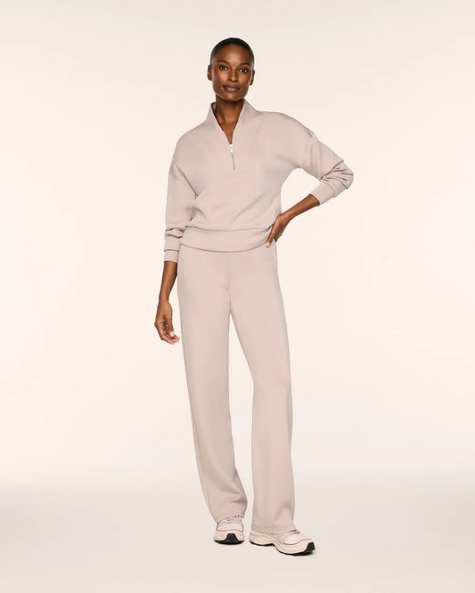 A model wearing a light beige two-piece tracksuit consisting of a long-sleeve quarter-zip pullover and matching wide-leg sweatpants, paired with white sneakers, against a solid off-white studio background.