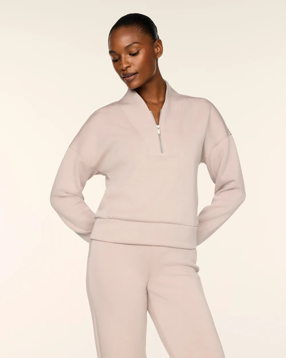 A model wearing a light beige matching lounge set consisting of a cropped quarter-zip sweatshirt and matching sweatpants, photographed against a solid off-white studio background.
