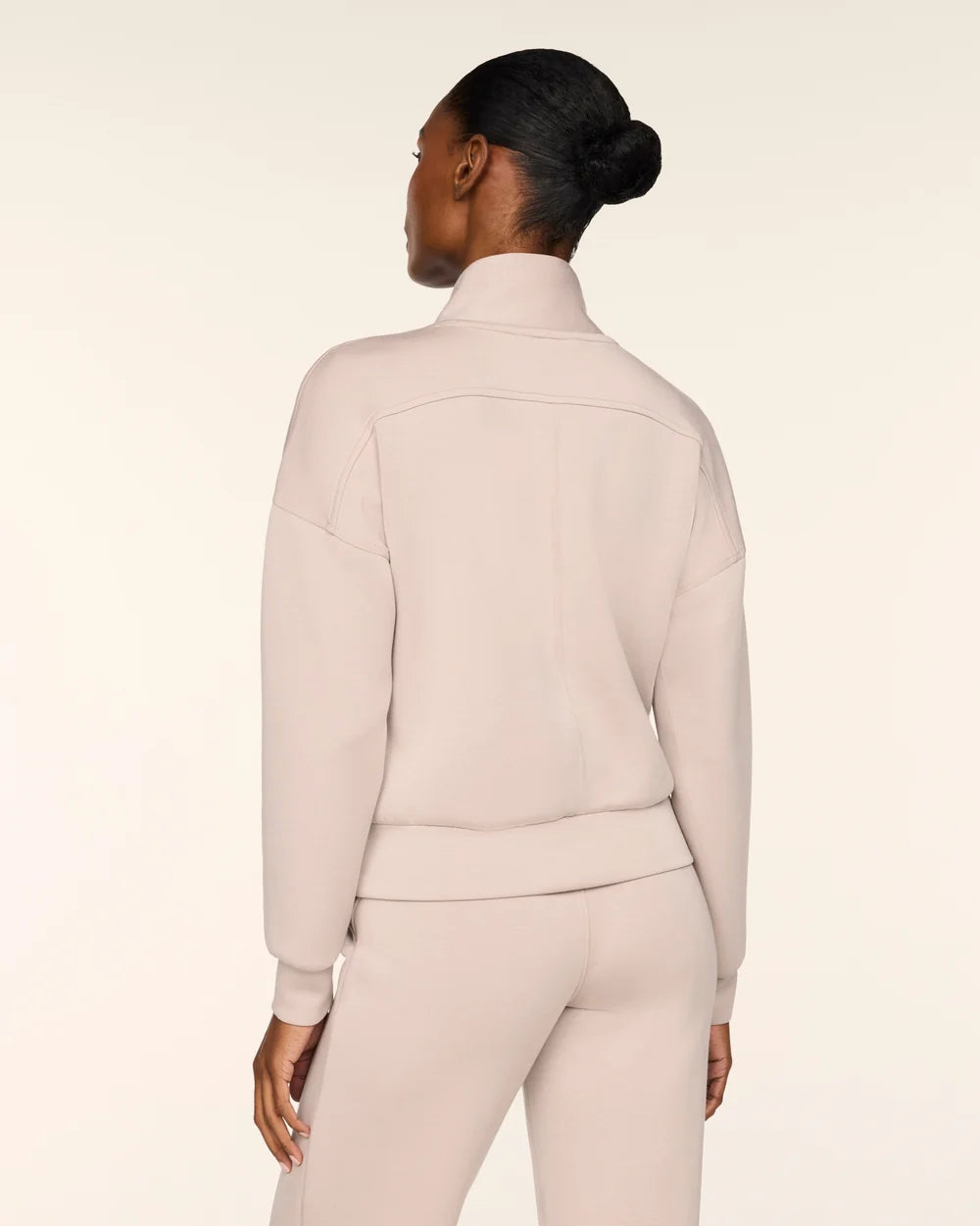 A model shown from the back wearing a matching light beige knit tracksuit, featuring a mock neck sweatshirt and coordinating pants, against a solid cream studio background.