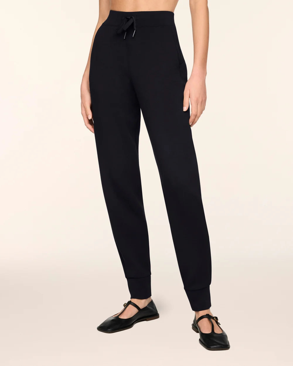 A model wearing black jogger-style pants with a drawstring waist and cuffed ankles, paired with black flat shoes, against a plain light beige studio background.